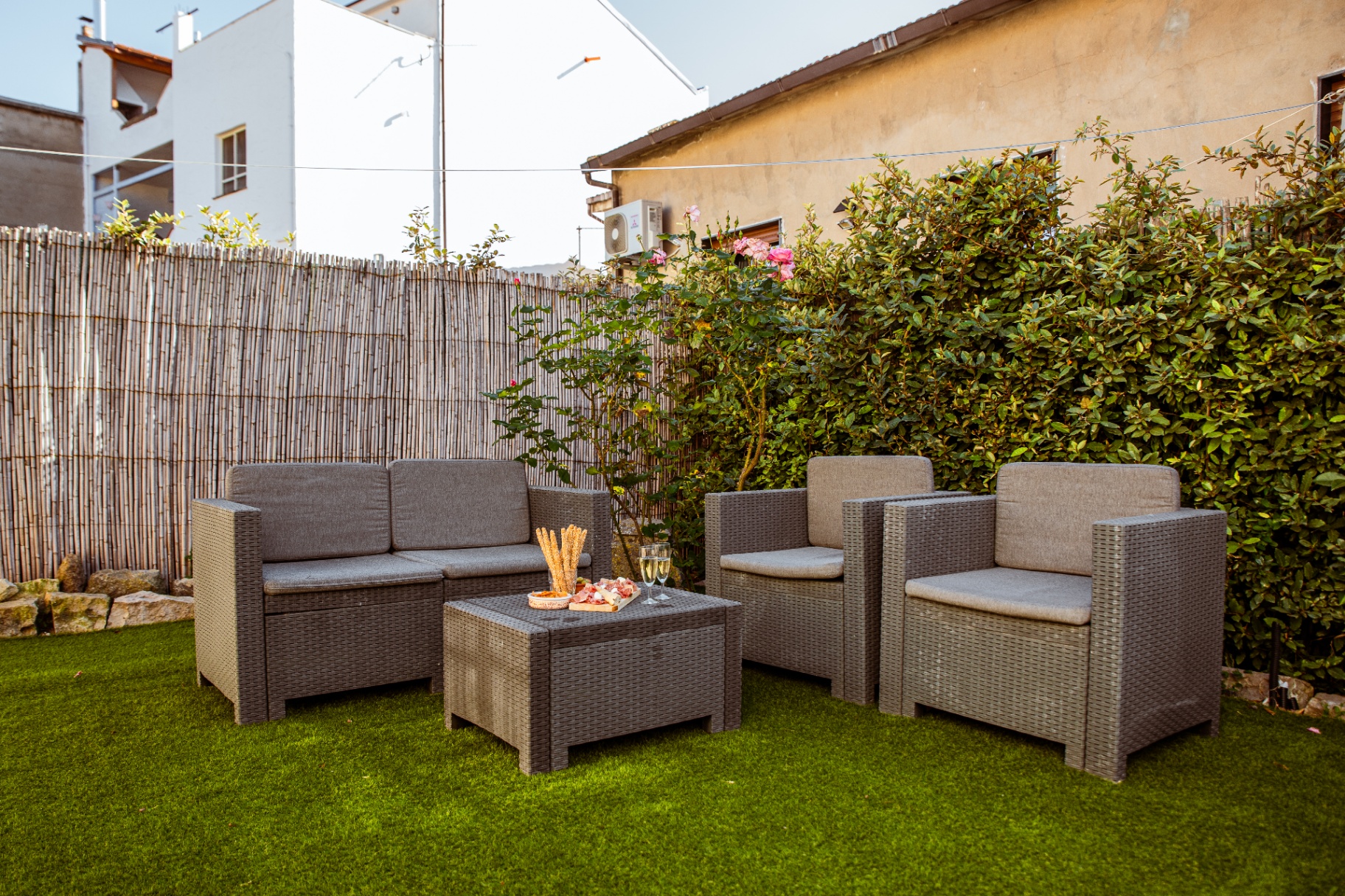 Salon de jardin en résine tressée sur la pelouse, rosiers en fleurs et haie de laurier